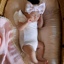 Baby in a white outfit with large pink bows lying on a pink cushion in a wicker basket.